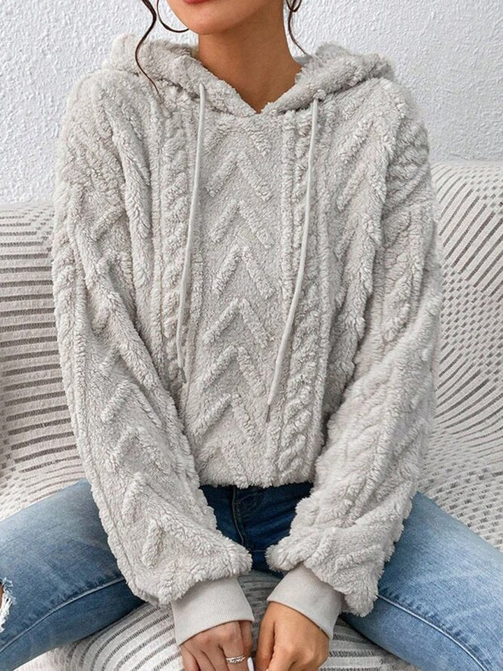 Woman sitting on a sofa in a light grey teddy hoodie with a chevron and cable pattern | Grace Aura