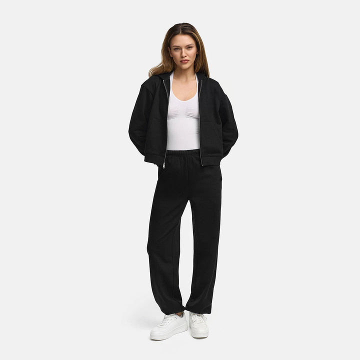 Woman wearing a black zip-up hoodie open over a white top with matching trousers, full length | Grace Aura