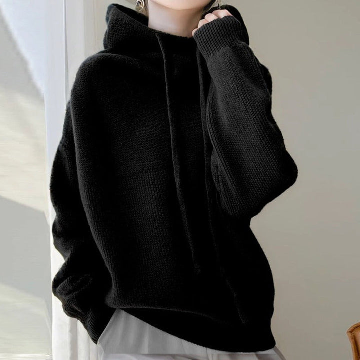 Woman wearing a black chunky-knit hoodie with ribbed cuffs, one hand at the collar | Grace Aura