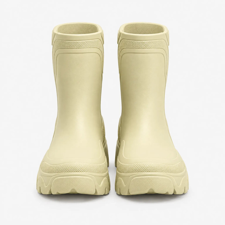 Front view of a pair of pale butter yellow chunky ankle height rubber rain boots with lugged soles on a plain light grey studio background.