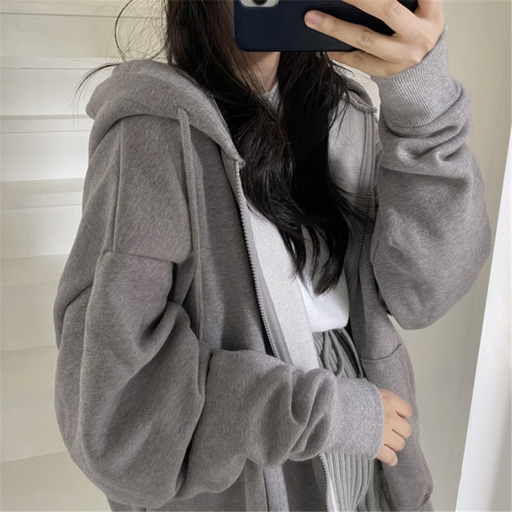 A woman takes a mirror selfie wearing an oversized grey fleece zip-up hoodie over a white top, with her face hidden behind a phone.