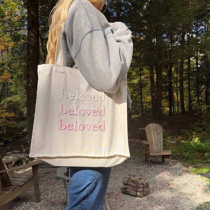Cream canvas tote bag with embroidered text, worn on woman&