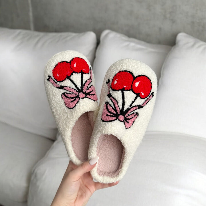 Cream plush slippers with cherry and pink bow embroidery, held by hand | Grace Aura