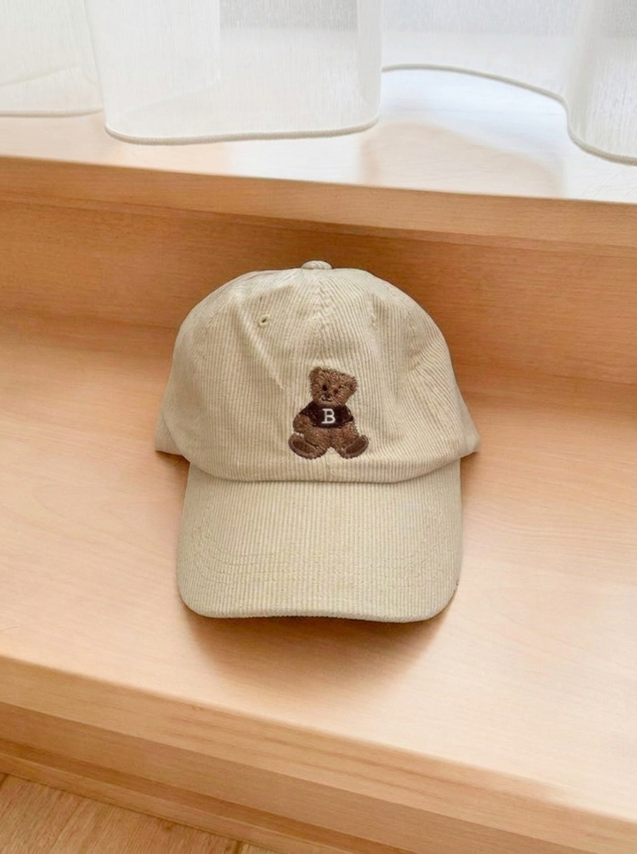 Beige corduroy baseball cap with embroidered teddy bear, laid flat on wood | Grace Aura