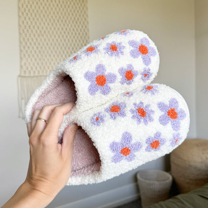 White plush slippers with purple and orange floral pattern, held by hand | Grace Aura