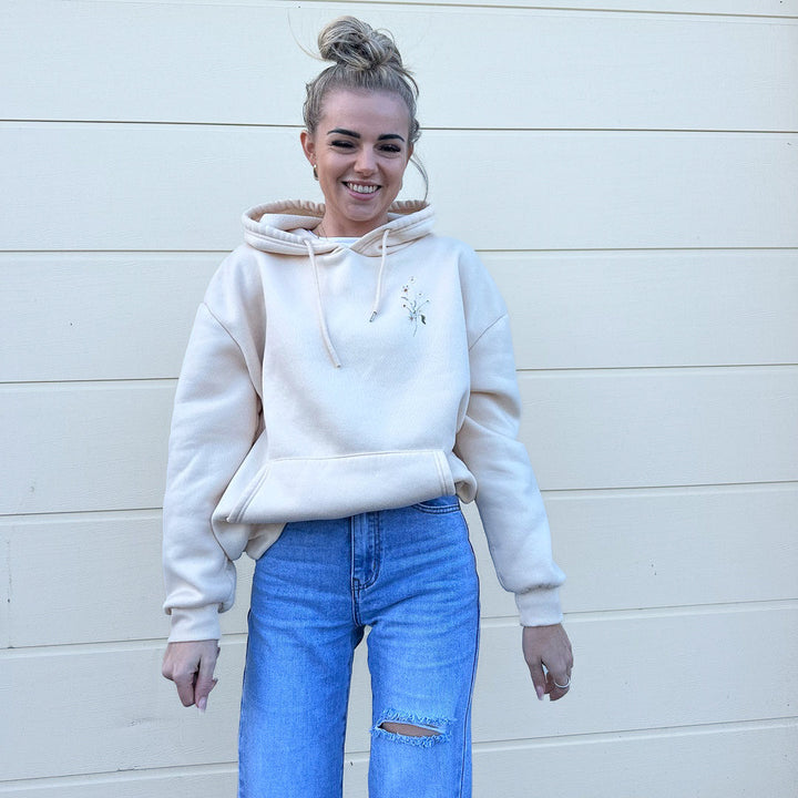 Woman wearing cream fleece hoodie with small floral embroidery and blue jeans | Grace Aura