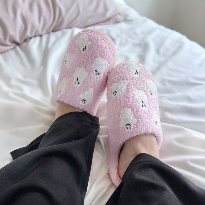 Pink fuzzy slippers with white ghost print, worn on feet, on bed | Grace Aura