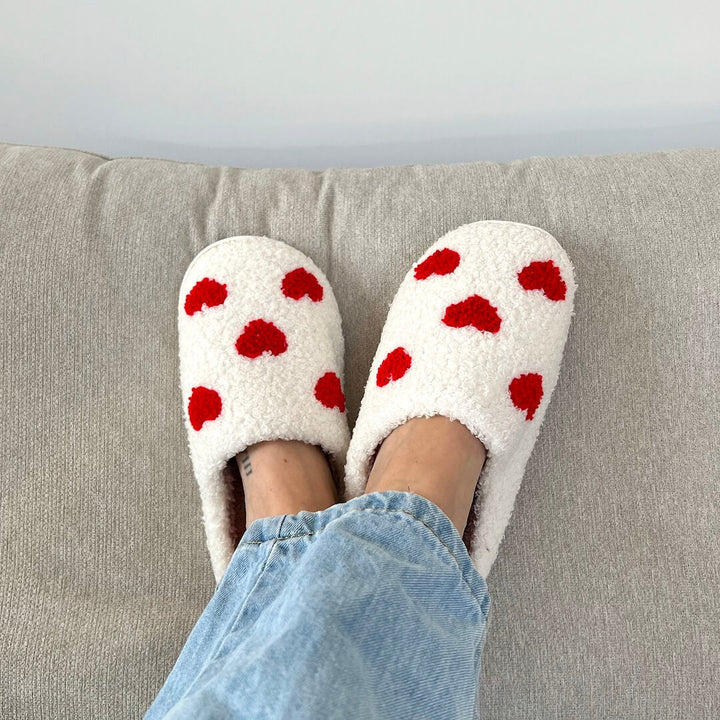 White plush slippers with red heart pattern, worn by person on couch | Grace Aura