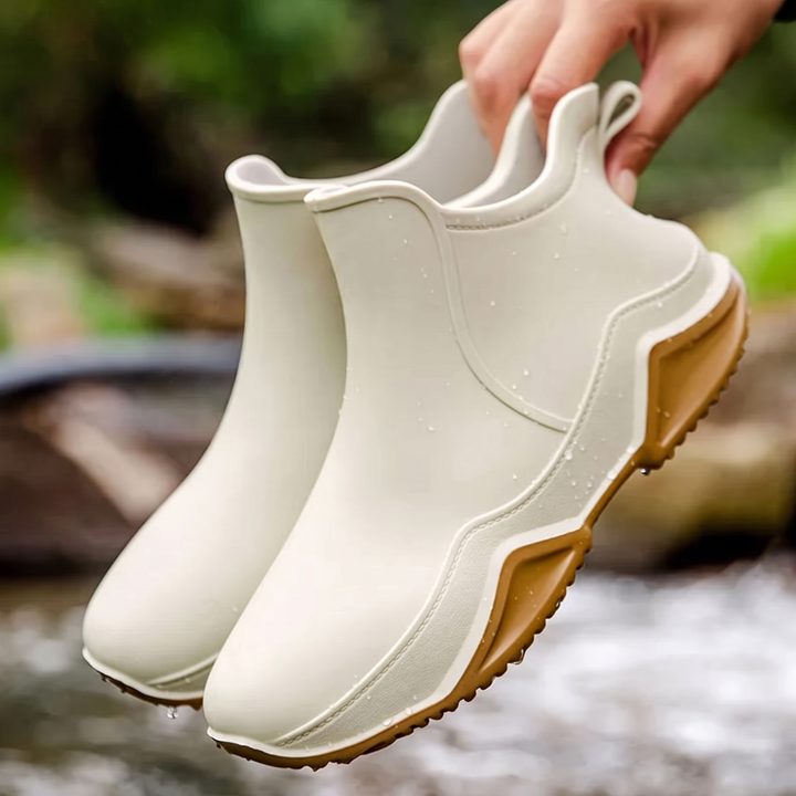 A hand holds a pair of cream ankle rain boots with tan treaded soles above a stream outdoors.