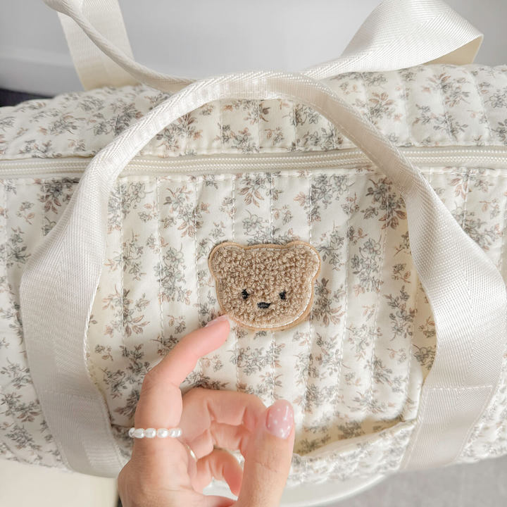 A close-up of a cream floral quilted duffle bag with a teddy bear patch on the front pocket, held open by a hand.
