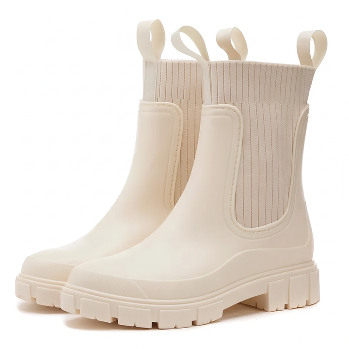 A pair of cream rubber Chelsea style rain boots with ribbed knit elastic side panels, pull loops and chunky lug soles, shown in a three quarter view on a white background.