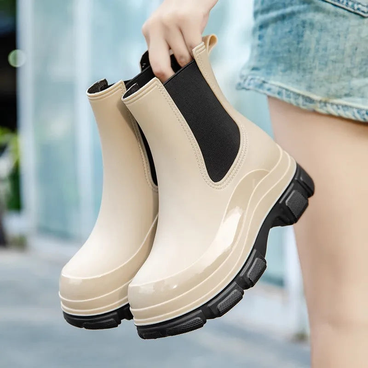 A hand holds a pair of glossy cream platform Chelsea rain boots with black elastic side panels and a black lug sole, beside a person in denim shorts on a blurred street.