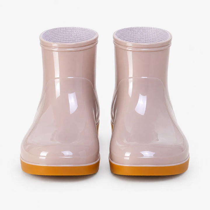 A pair of glossy pale pink ankle rain boots with mustard orange soles, shown from the front on a plain white studio background.