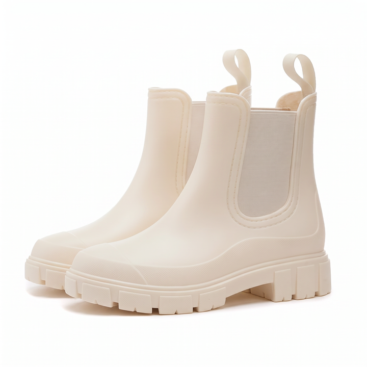 Studio shot of a pair of cream ankle rain boots in Chelsea style, with beige elastic side panels, pull tabs and chunky lugged soles, seen in three quarter view on a white background; the side panel carries a small woven tab reading UYIRAIN, which appears to be an anonymous supplier label.