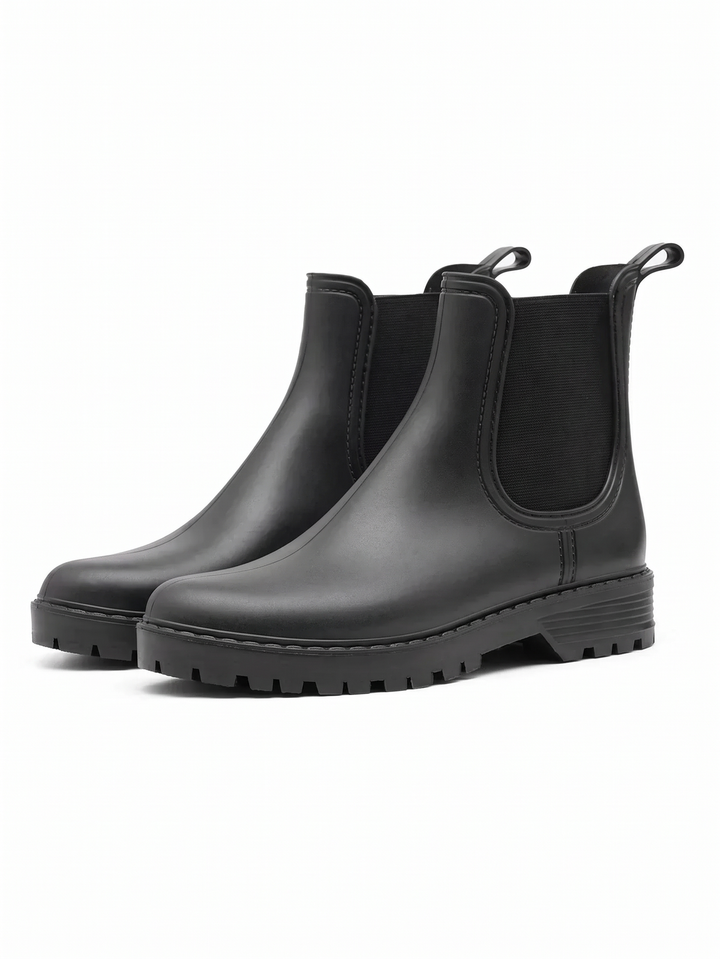 A pair of matte black Chelsea rain boots with black elastic side panels, rear pull tabs and chunky lug soles, shown three quarter on a white background.