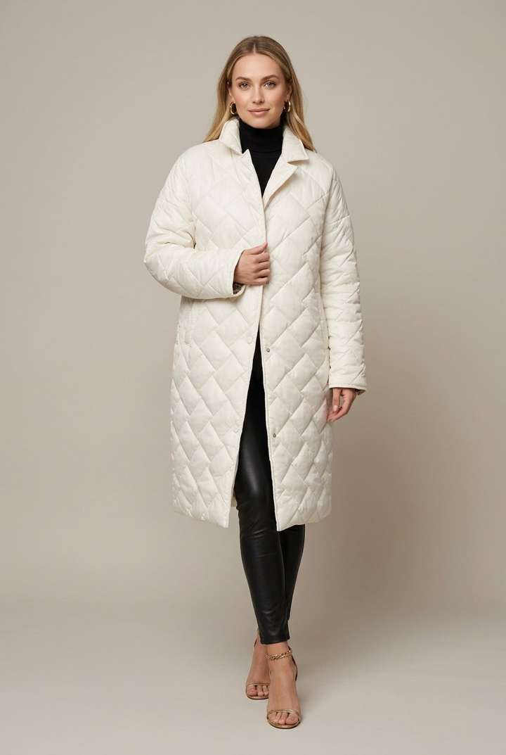 A woman wears a long cream quilted puffer coat over a black turtleneck and black leather pants, standing in a studio.