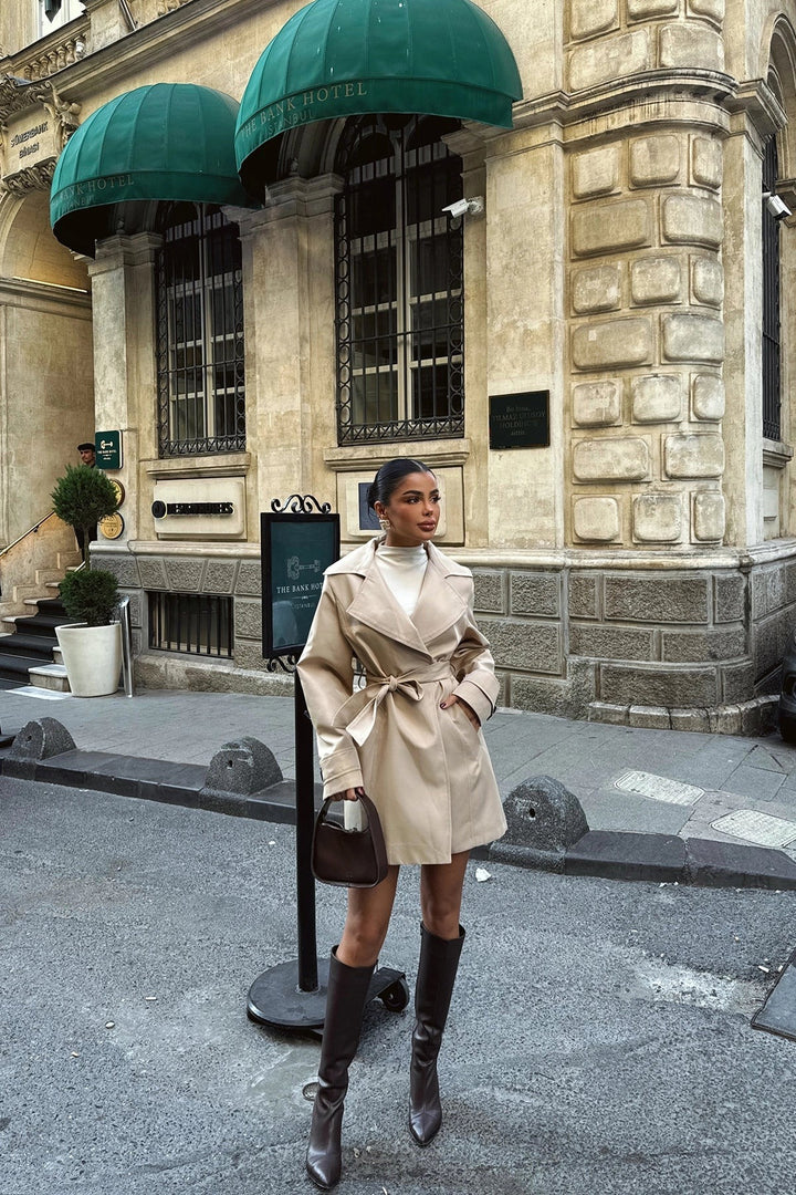 Woman in a short beige trench coat with tie belt, worn with knee-high boots on a city street