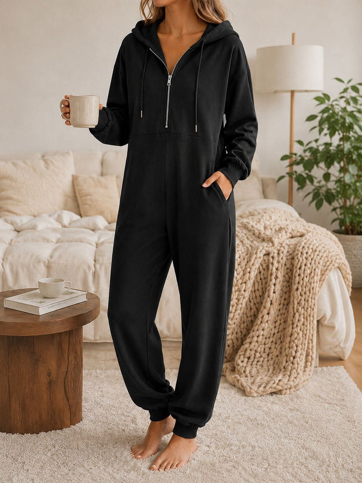Black hooded onesie with a half zip and side pockets, front view in a bedroom, holding a mug