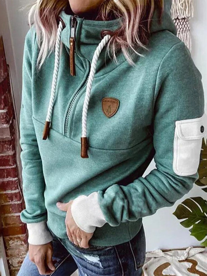 Woman wearing a mint green half-zip hoodie with a high collar, white sleeve pocket and brown patch, one hand in her jeans pocket | Grace Aura
