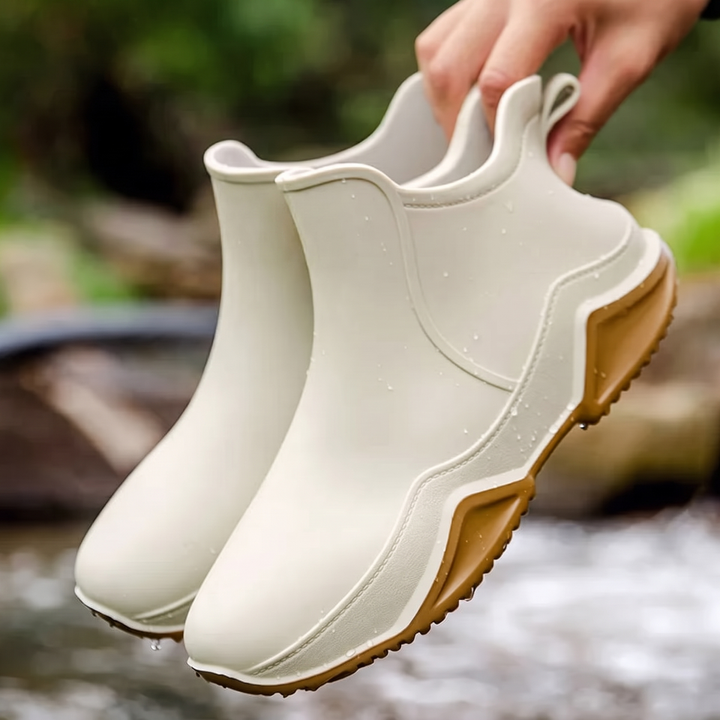A hand holds a pair of cream slip-on rain boots with tan outsoles and water droplets, outdoors above a blurred stream.