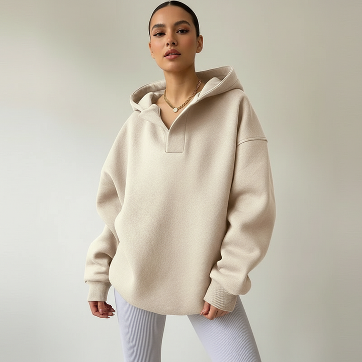 A woman in a studio poses facing the camera wearing an oversized cream hoodie with a notched V neckline, a gold chain necklace and white ribbed leggings against a plain light grey background.