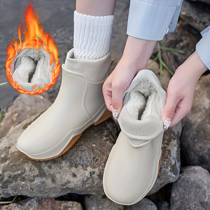 A woman kneels on rocks by water pulling plush-lined cream rain boots onto her feet, with a flame graphic highlighting the fleece lining of one boot.