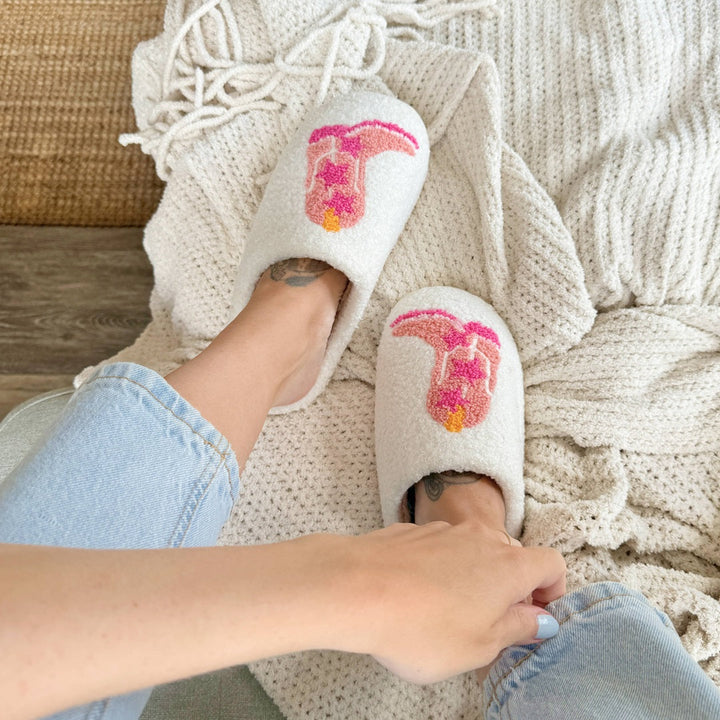 Cream slippers with pink cowboy boot embroidery, worn on feet, cozy blanket background | Grace Aura