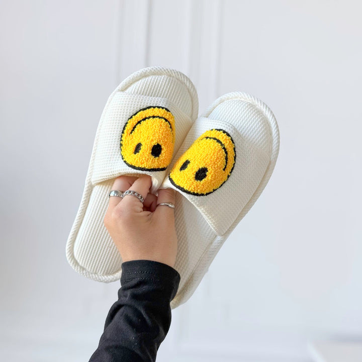 White terry slippers with yellow smiley face patches, held by hand | Grace Aura