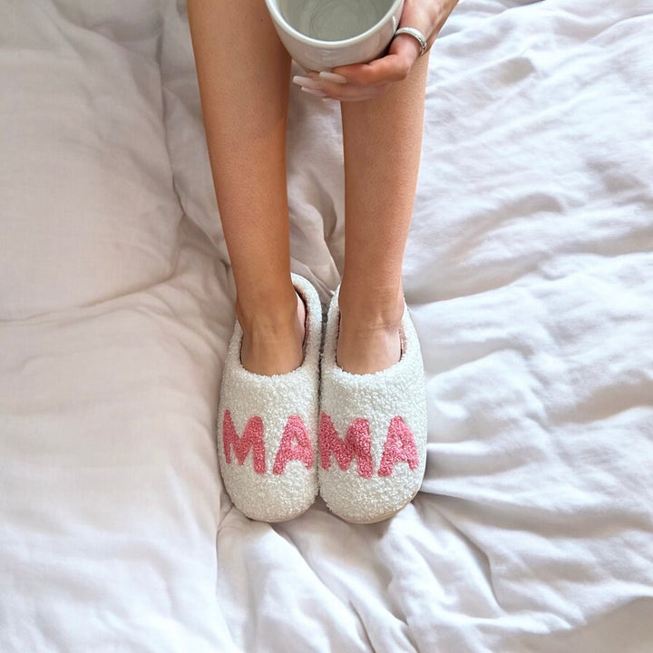 White fuzzy slippers with pink lettering, worn by person on bed | Grace Aura