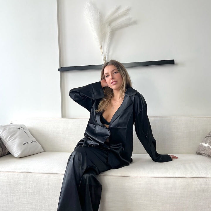 Woman wearing black satin pyjama set, seated on cream sofa indoors | Grace Aura