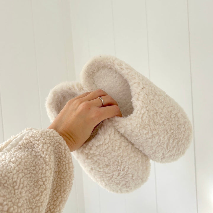 Cream sherpa slip-on slippers held by hand in fuzzy sleeve | Grace Aura