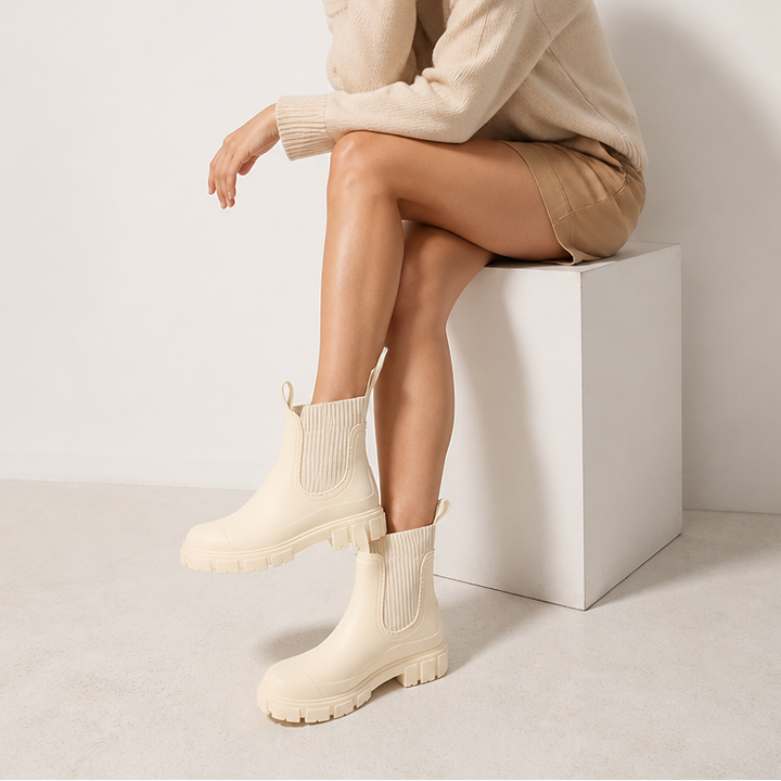 A woman sits on a white block wearing cream chelsea style rain boots with ribbed elastic collars.