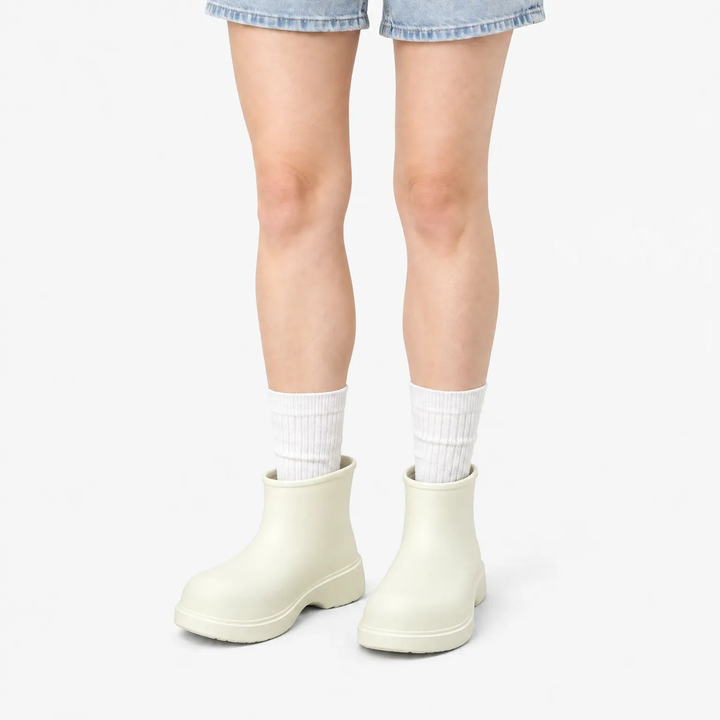 A person wearing white socks and cream colored ankle rain boots with a chunky sole, shown from the front cropped at the thighs.