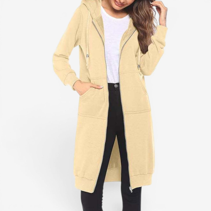 A woman in a studio wears a long beige full-zip hoodie, open over a white t-shirt and black jeans, with one hand in a front pocket.