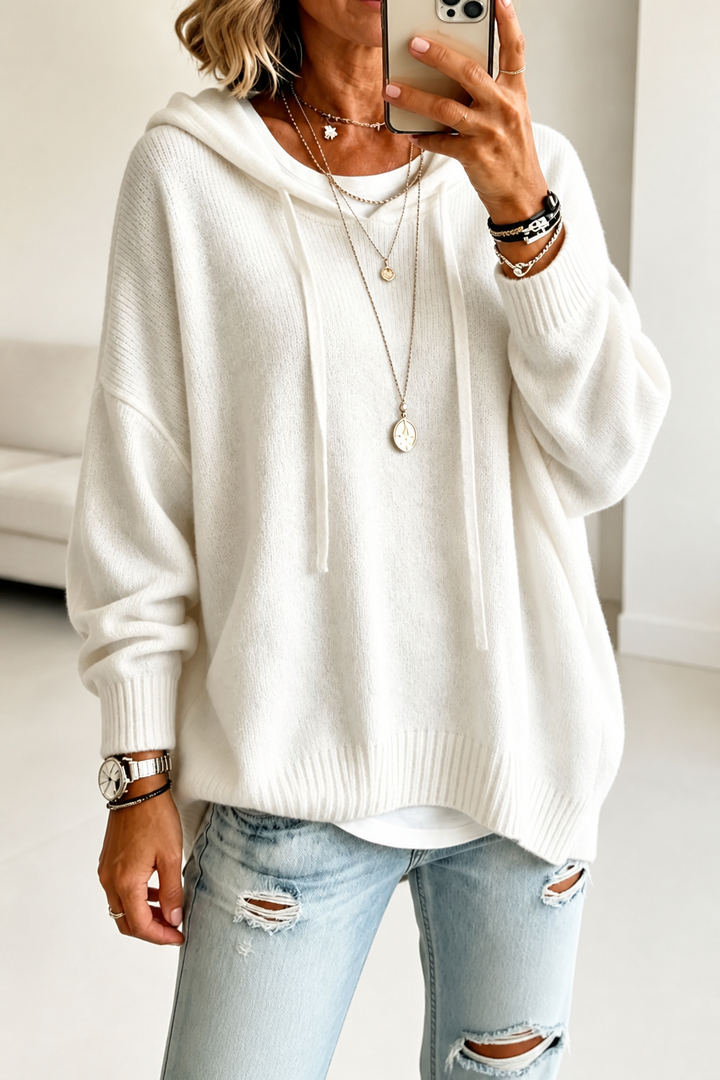 A woman takes a mirror selfie in a bright living room wearing an oversized white knit hoodie with drawstrings over a white tee and light ripped jeans, with layered necklaces and bracelets.