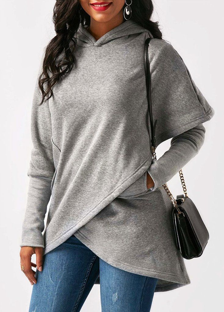 A woman in a studio wears a grey asymmetric wrap front longline hoodie with jeans, carrying a black chain strap shoulder bag.