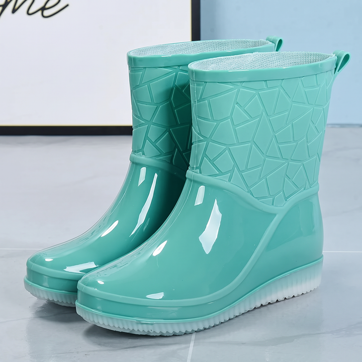 A pair of mint green mid-calf rain boots with a geometric patterned upper, glossy lower and translucent sole, shown at a three quarter angle on a grey floor.