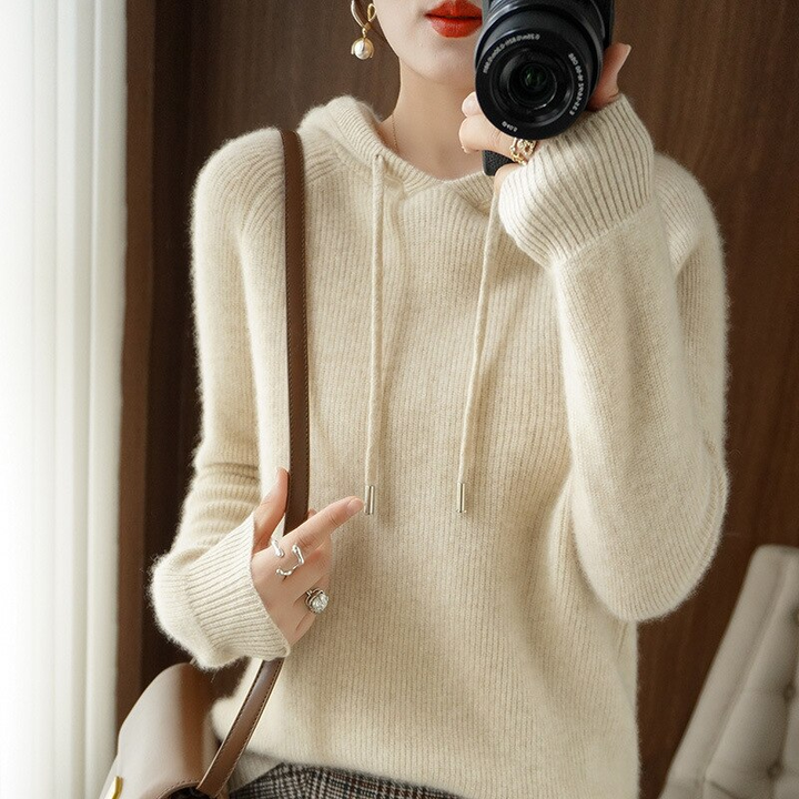 A woman in a cream ribbed knit hoodie with drawstrings stands in a room holding a camera in front of most of her face, with a brown shoulder bag strap across her body.