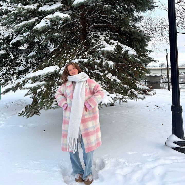 Woman wearing pink plaid oversized coat with white scarf, standing outdoors in snow | Grace Aura